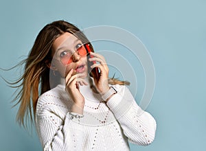 Adolescent in orange sunglasses, watch, bracelet and sweater. She talking by phone, surprised, posing on blue background. Close up