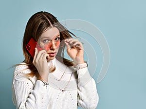 Adolescent in orange sunglasses, watch, bracelet and sweater. She talking by phone, surprised, posing on blue background. Close up