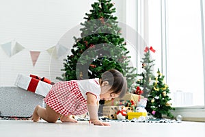 Adorable 8 months Asian baby girl having fun crawling on the floor in front of a Christmas tree, among gift boxes, dolls and toys