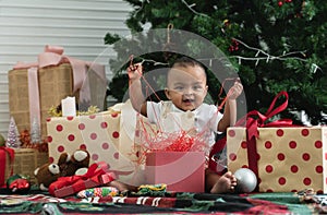 Adorable 9 months African baby smiling and having fun to open gift box, sitting in front of Christmas tree on floor
