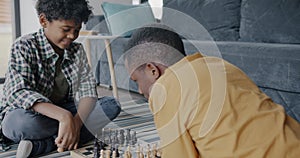Adorable African American child playing chess with loving dad thinking and moving figures on chessboard on floor at home