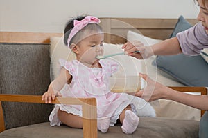 Adorable Asian baby eating food from spoon, mother feeding daughter puree or solid foods, sitting on chair at home. Little cute