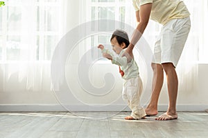 Adorable Asian baby toddler learning to walk with dad helping at home. Little baby boy looking in front to walking step by step