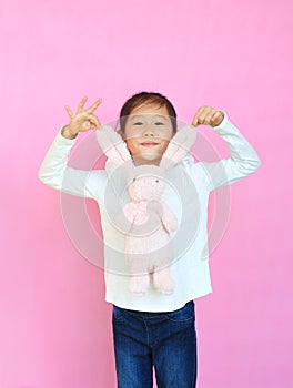 Adorable asian kid girl holding ears of pink rabbit doll isolated on pink background. Happy child on Easter