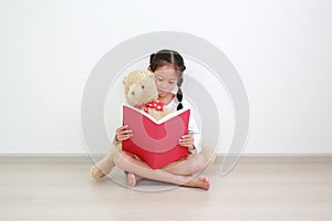 Adorable asian little child girl reading a book with hugging a teddy bear doll sitting against white wall in the room