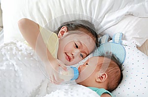 Adorable Asian older sister feeding milk from bottle for newborn baby lying on the bed