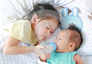 Adorable Asian older sister feeding milk from bottle for newborn baby lying on the bed