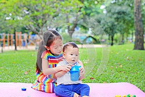 Adorable Asian sister sitting on pink mattress mat take care her little brother to drinking water from Baby sippy cup with straw