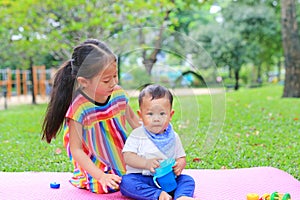 Adorable Asian sister sitting on pink mattress mat take care her little brother to drinking water from Baby sippy cup with straw