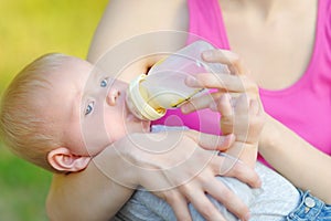 Adorable baby boy drinking milk from bottle in mother hands. Mom feeding child fo artificial nutrition formula