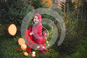 Adorable baby boy in the red knit jumpsuit and Santa hat  and holding rocking-horse with pine trees on the background