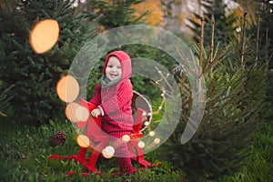 Adorable baby boy in the red knit jumpsuit and Santa hat  and holding rocking-horse with pine trees on the background