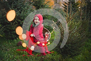 Adorable baby boy in the red knit jumpsuit and Santa hat  and holding rocking-horse with pine trees on the background