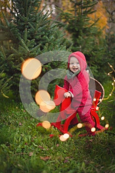 Adorable baby boy in the red knit jumpsuit and Santa hat  and holding rocking-horse with pine trees on the background