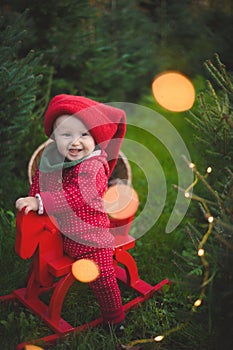 Adorable baby boy in the red knit jumpsuit and Santa hat  and holding rocking-horse with pine trees on the background