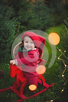 Adorable baby boy in the red knit jumpsuit and Santa hat  and holding rocking-horse with pine trees on the background