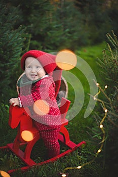 Adorable baby boy in the red knit jumpsuit and Santa hat  and holding rocking-horse with pine trees on the background