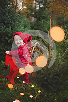 Adorable baby boy in the red knit jumpsuit and Santa hat  and holding rocking-horse with pine trees on the background