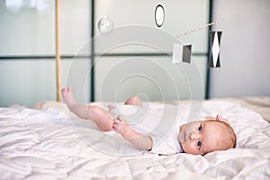 Adorable baby boy in white sunny bedroom lying and looks at Munari Montessori mobile.