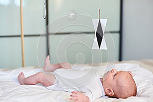 Adorable baby boy in white sunny bedroom lying and looks at Munari Montessori mobile.