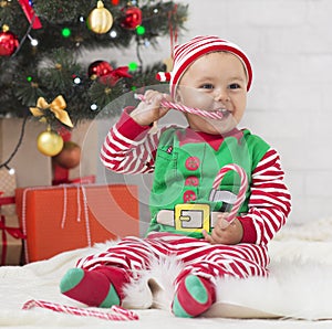 Adorable baby elf eating Christmas lollipop under Xmas tree