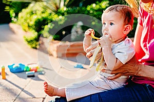 Adorable baby with funny face while trying to bite a banana to feed, in the arms of his mother