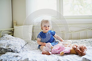 Adorable baby girl in blue dress sitting on bed and playing with a doll