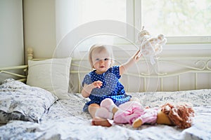 Adorable baby girl in blue dress sitting on bed and playing with doll, teddy bear and dog