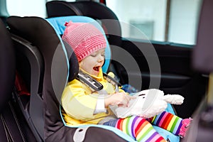 Adorable baby girl with blue eyes and in colorful clothes sitting in car seat. Toddler child in winter clothes going on