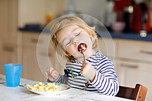 Adorable baby girl eating from spoon noodle, pasta macaroni. Cute healthy toddler child, daughter with spoon sitting in