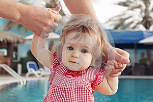 Adorable baby girl holding father`s hands while learning to walk outdoors. Swimming pool background. Baby looking at