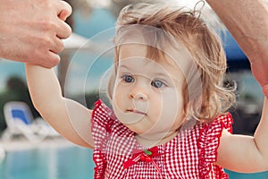 Adorable baby girl holding father`s hands while learning to walk outdoors. Swimming pool background. Baby looking up.