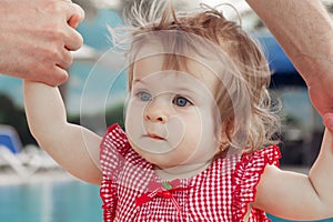Adorable baby girl holding father`s hands while learning to walk outdoors. Swimming pool background. Baby looking up.