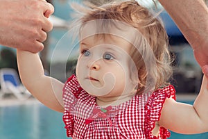 Adorable baby girl holding father`s hands while learning to walk outdoors. Swimming pool background. Baby looking up.