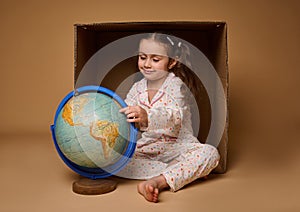 Adorable baby girl in pajamas sitting ahead a cardboard box and examining the Earth globe,  over beige background with