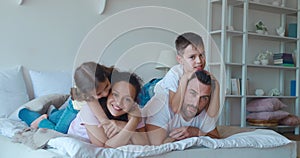 Adorable baby girl tenderlykissing and hugging her African American mother, lying down on the bed next to her brother on