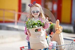 A little girl with a big trolley buys fresh food and vegetables in a big supermarket in the summer alone.