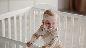 Adorable Baby With Pacifier In Mouth Standing In Crib And Waving Hand