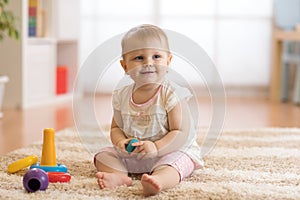 Adorable baby playing with colorful rainbow toy pyramid sitting on rug in white sunny bedroom. Toys for little kids