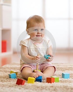Adorable baby playing with colorful toy pyramid sitting on carpet in white sunny bedroom. Toys for little kids. Child