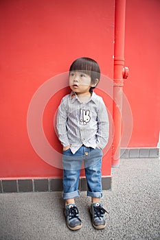 Adorable baby in a red shirt and jeans holding a hose standing in front of a red wall