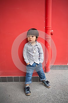 Adorable baby in a red shirt and jeans holding a hose standing in front of a red wall