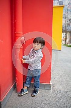 Adorable baby in a red shirt and jeans holding a hose standing in front of a red wall