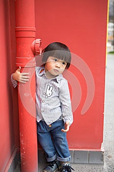 Adorable baby in a red shirt and jeans holding a hose standing in front of a red wall