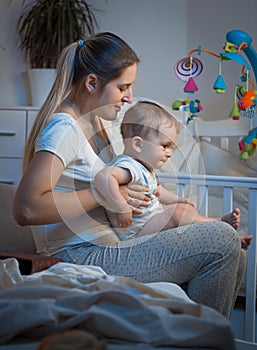 Adorable baby sitting on mothers lap in bedroom at night