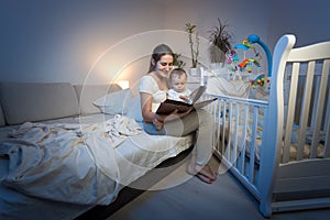 Adorable baby sitting on mothers lap and reading book before going to sleep