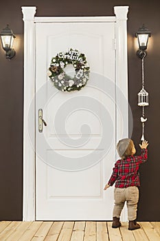 Adorable baby stands at the front door with a christmas wreath