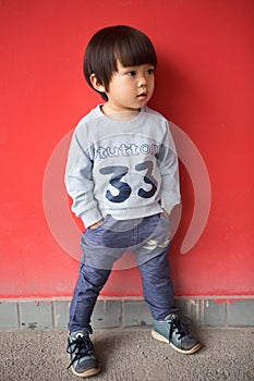 Adorable baby in stylish clothes posing in front of red wall