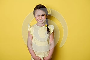 Adorable beautiful noble little child girl wearing yellow dress and roses in hairstyle, cutely smiles looking at camera