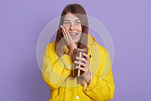 Adorable beautiful young woman holding thermo cup of tea and keeping palm on cheek, keeps mouth opened, looks excited, wearing